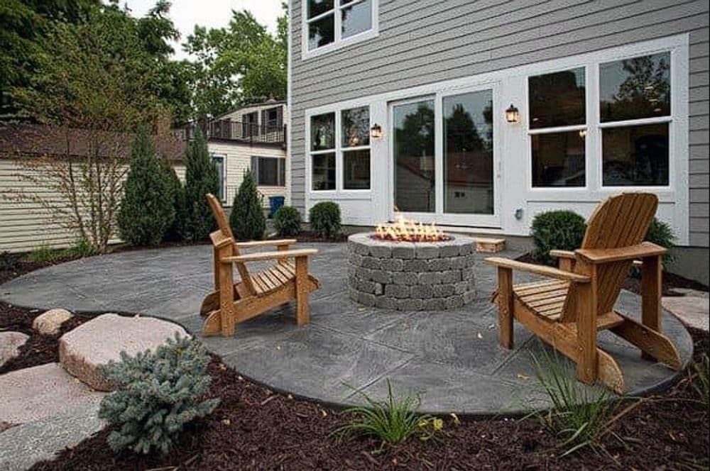 Outdoor patio with stone fire pit, wooden chairs, and landscaped greenery. Ideal for gatherings.